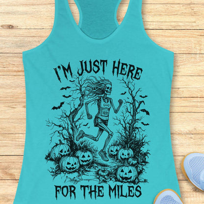 A turquoise racerback tank top laid flat on a light wood background, featuring a black graphic of a skeleton runner surrounded by pumpkins and bats with the text 'I'M JUST HERE FOR THE MILES'. A partial white sneaker is visible at the bottom right.