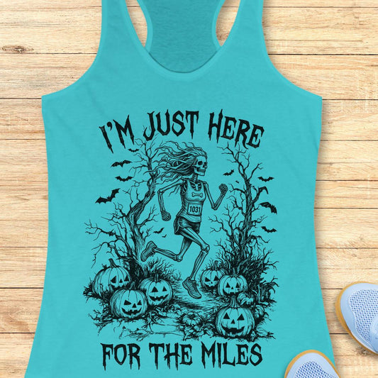A turquoise racerback tank top laid flat on a light wood background, featuring a black graphic of a skeleton runner surrounded by pumpkins and bats with the text 'I'M JUST HERE FOR THE MILES'. A partial white sneaker is visible at the bottom right.