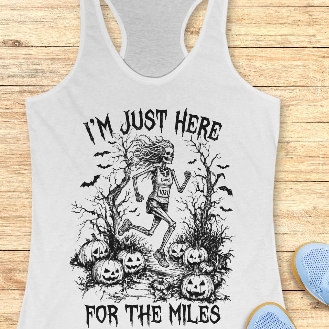 A white racerback tank top with a black graphic of a skeleton runner surrounded by pumpkins and bats, with the text 'I'M JUST HERE FOR THE MILES', shown on a wooden background with blue sneakers partially visible.