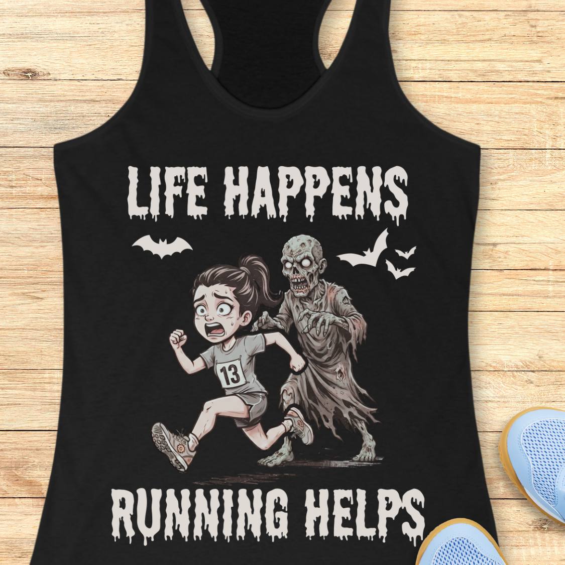 A black racerback tank top laid flat on a light wood surface, featuring a Halloween-themed graphic with the text 'LIFE HAPPENS RUNNING HELPS' and an illustration of a girl running alongside a zombie. Partial blue shoes are visible at the bottom right.