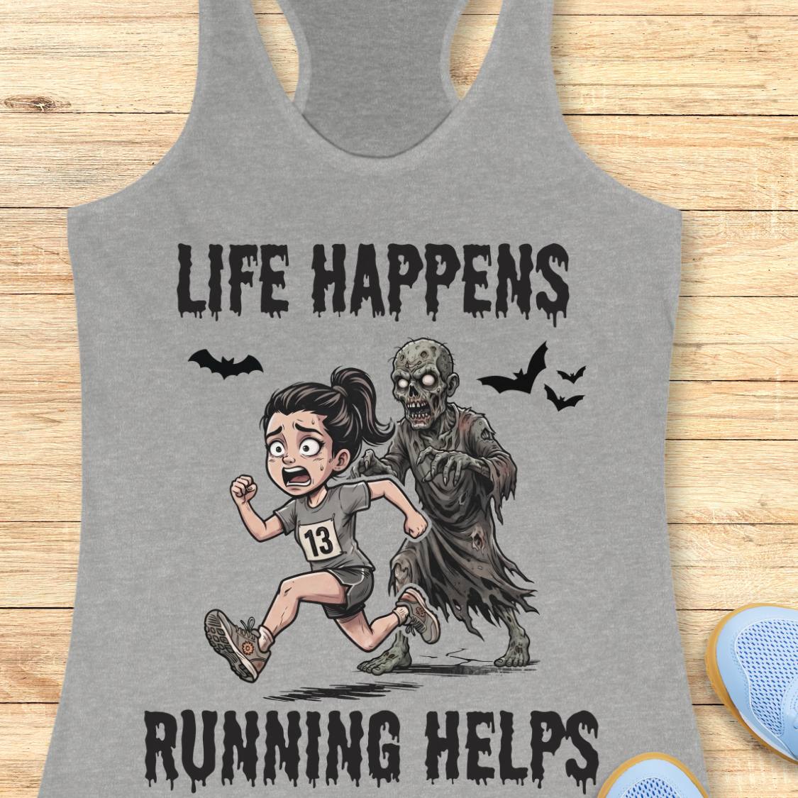 A heather gray racerback tank top laid flat on a light wood surface, featuring a Halloween graphic of a girl running with a zombie and the text 'LIFE HAPPENS RUNNING HELPS'.