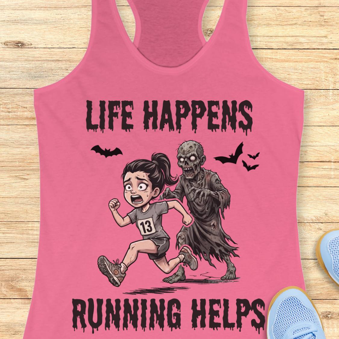 A pink racerback tank top laid flat on a light wood surface, featuring a cartoon graphic of a woman running while being chased by a zombie, with the text 'LIFE HAPPENS RUNNING HELPS' and small bat silhouettes.