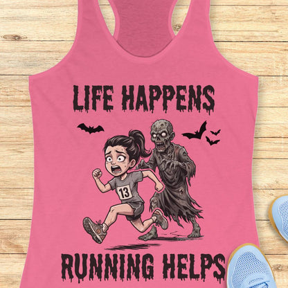 A pink racerback tank top laid flat on a light wood surface, featuring a cartoon graphic of a woman running while being chased by a zombie, with the text 'LIFE HAPPENS RUNNING HELPS' and small bat silhouettes.