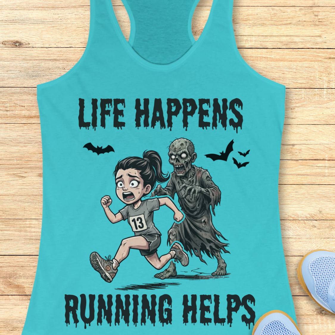 A turquoise racerback tank top laid flat on a light wood surface, featuring a cartoon graphic of a girl running alongside a zombie with the text 'LIFE HAPPENS RUNNING HELPS'.