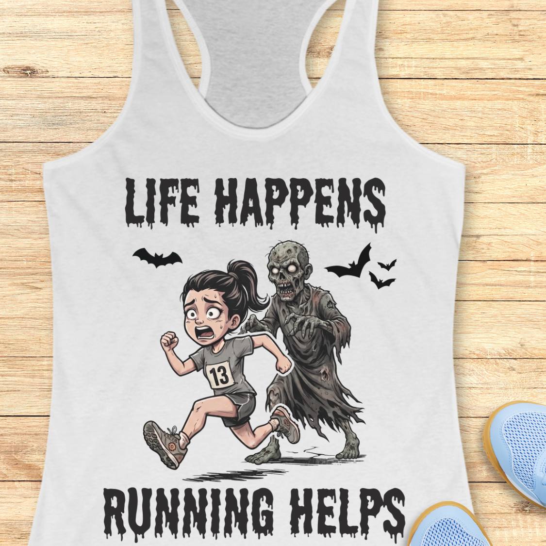 A white racerback tank top laid flat on a light wood surface, featuring a cartoon graphic of a girl running beside a zombie with the text 'LIFE HAPPENS RUNNING HELPS'. Partial blue shoes are visible at the bottom right as styling props.