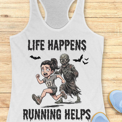 A white racerback tank top laid flat on a light wood surface, featuring a cartoon graphic of a girl running beside a zombie with the text 'LIFE HAPPENS RUNNING HELPS'. Partial blue shoes are visible at the bottom right as styling props.