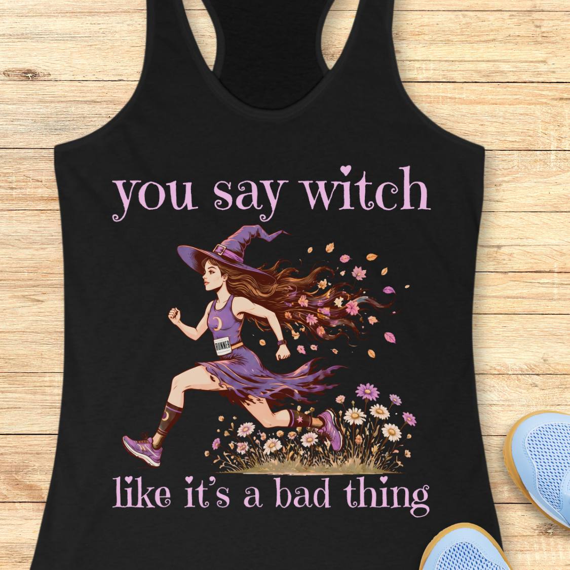 A black racerback tank top laid flat on a light wood surface, featuring a graphic of a witch running through flowers with the text 'you say witch like it's a bad thing' in lavender. Part of a blue shoe is visible at the bottom right as a styling prop.