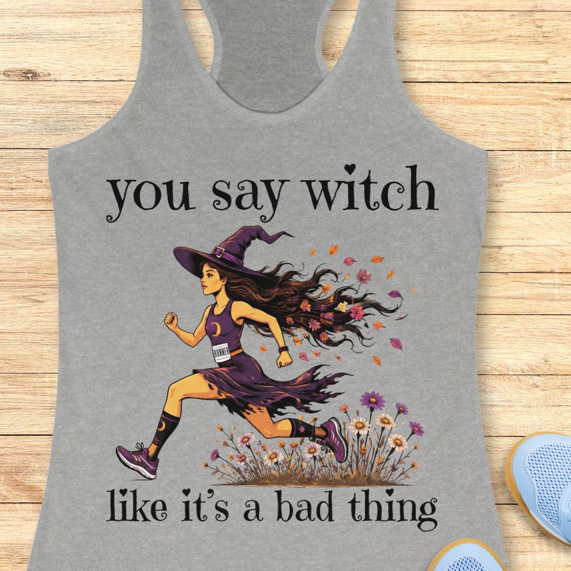 A heather gray racerback tank top laid flat on a light wood surface, featuring a graphic of a running witch with the text 'you say witch like it's a bad thing'.