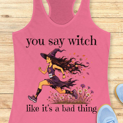 A pink racerback tank top laid flat on a light wood surface, featuring a printed illustration of a witch running with the text 'you say witch like it's a bad thing.' A small portion of a blue shoe is visible at the lower right edge.