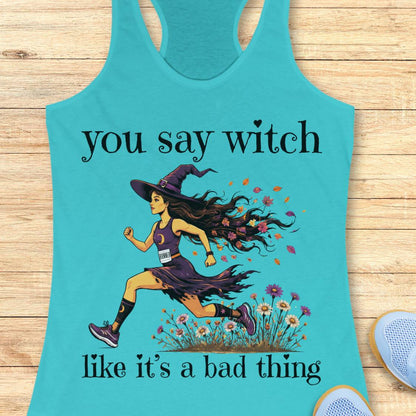 A turquoise racerback tank top laid flat on a light wood surface, featuring a printed graphic of a witch running through flowers with the text 'you say witch like it's a bad thing'. Part of a blue shoe is visible at the bottom right.