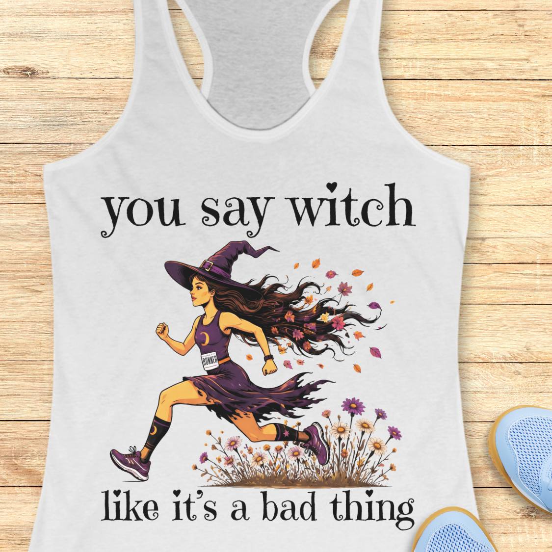 A white racerback tank top with a graphic print of a witch running, with the text 'you say witch like it's a bad thing'. The tank top is displayed flat on a light wood background with blue sneakers partially visible in the corner.