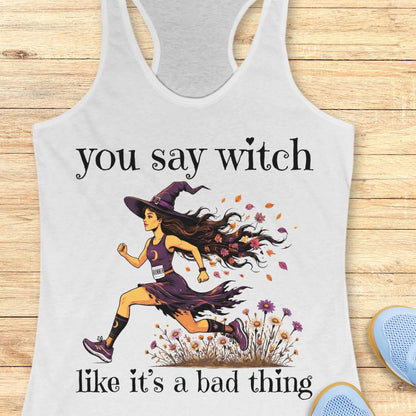 A white racerback tank top with a graphic print of a witch running, with the text 'you say witch like it's a bad thing'. The tank top is displayed flat on a light wood background with blue sneakers partially visible in the corner.