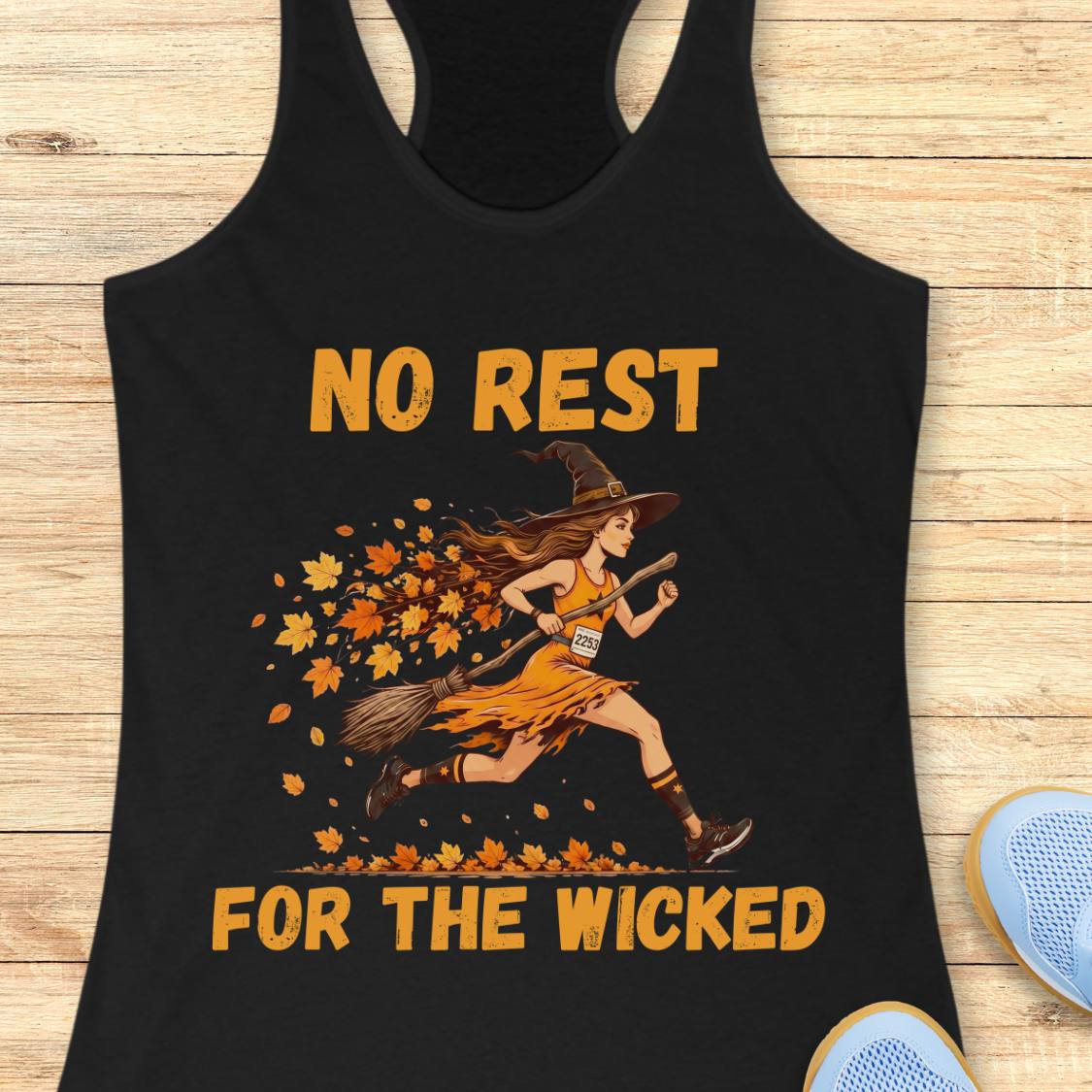 A black racerback tank top laid flat on a light wood surface, featuring a graphic of a witch running with a broom and falling orange leaves, with the text 'NO REST FOR THE WICKED' in orange. Partial blue shoes are visible at the bottom right as styling props.