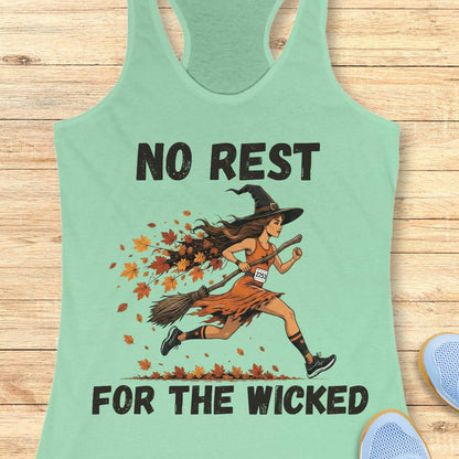 A mint green racerback tank top with a graphic of a witch running on a broomstick surrounded by autumn leaves, with the text 'NO REST FOR THE WICKED' printed in black. The tank top is displayed flat on a light wood background with partial blue shoes visible in the corners.