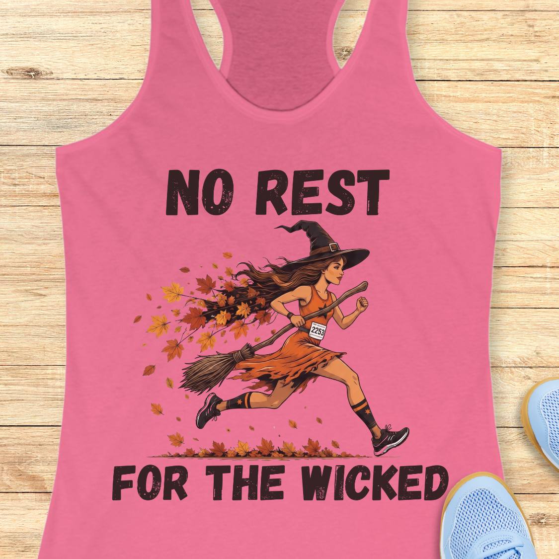 A pink racerback tank top laid flat on a light wood surface, featuring a graphic of a witch running while sweeping up falling autumn leaves, with the text 'NO REST FOR THE WICKED' in black. Partial blue shoes are visible at the bottom right as styling props.
