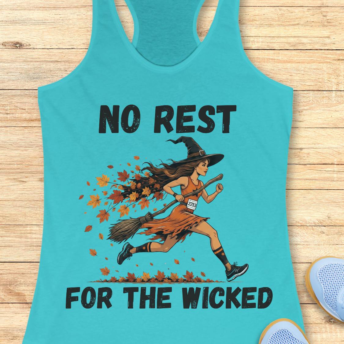 A turquoise racerback tank top laid flat on a light wood surface, featuring a graphic of a witch running with a broom and the text 'NO REST FOR THE WICKED' in black. Part of a blue shoe is visible at the bottom right.