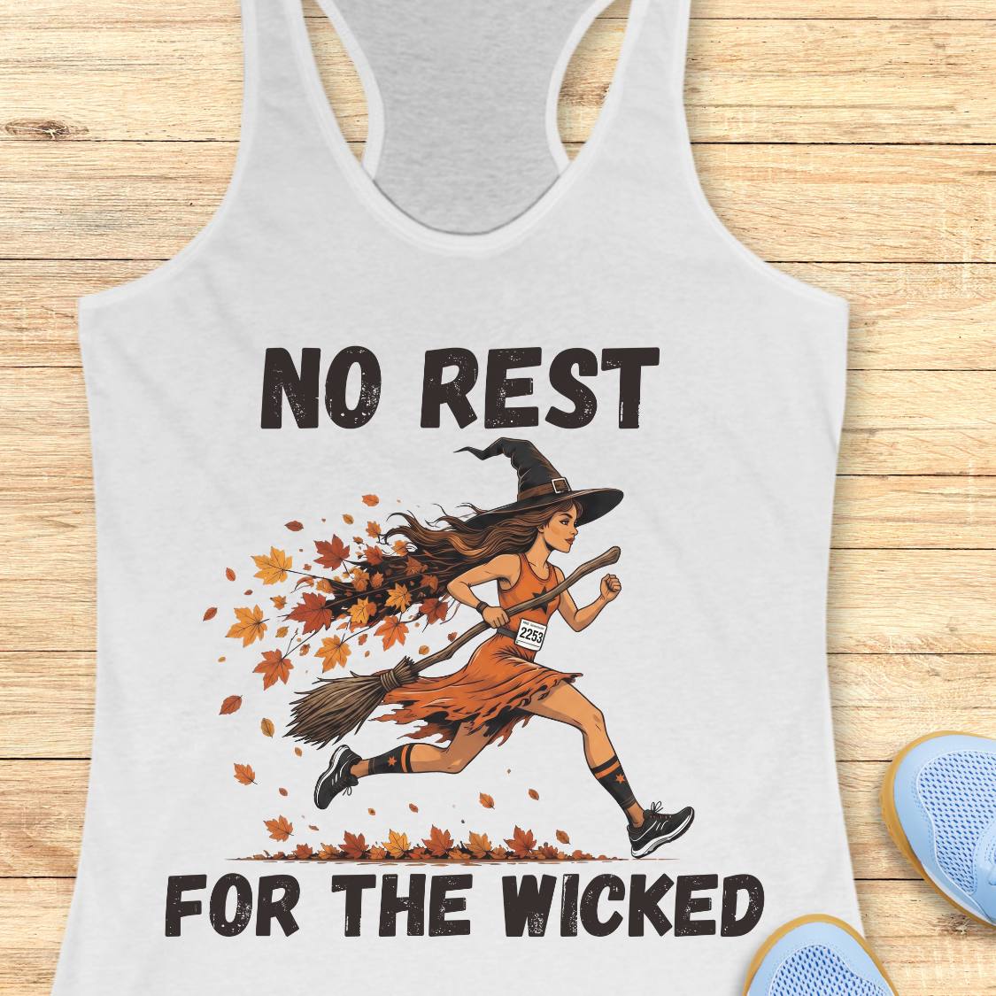 A white racerback tank top laid flat on a light wood surface, featuring a Halloween graphic of a witch running on a broomstick with falling autumn leaves. The text reads 'NO REST FOR THE WICKED' in black.