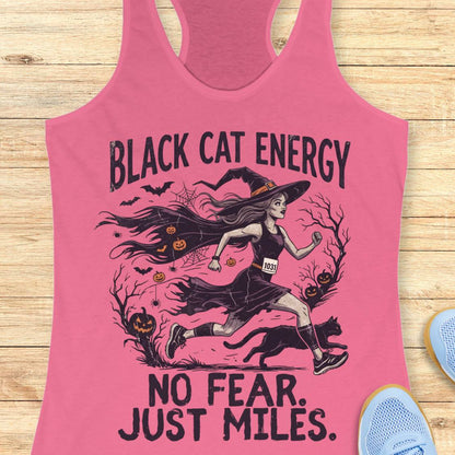 A pink racerback tank top laid flat on a light wood surface, featuring a Halloween-themed graphic of a witch running with a black cat and the text 'BLACK CAT ENERGY NO FEAR. JUST MILES.