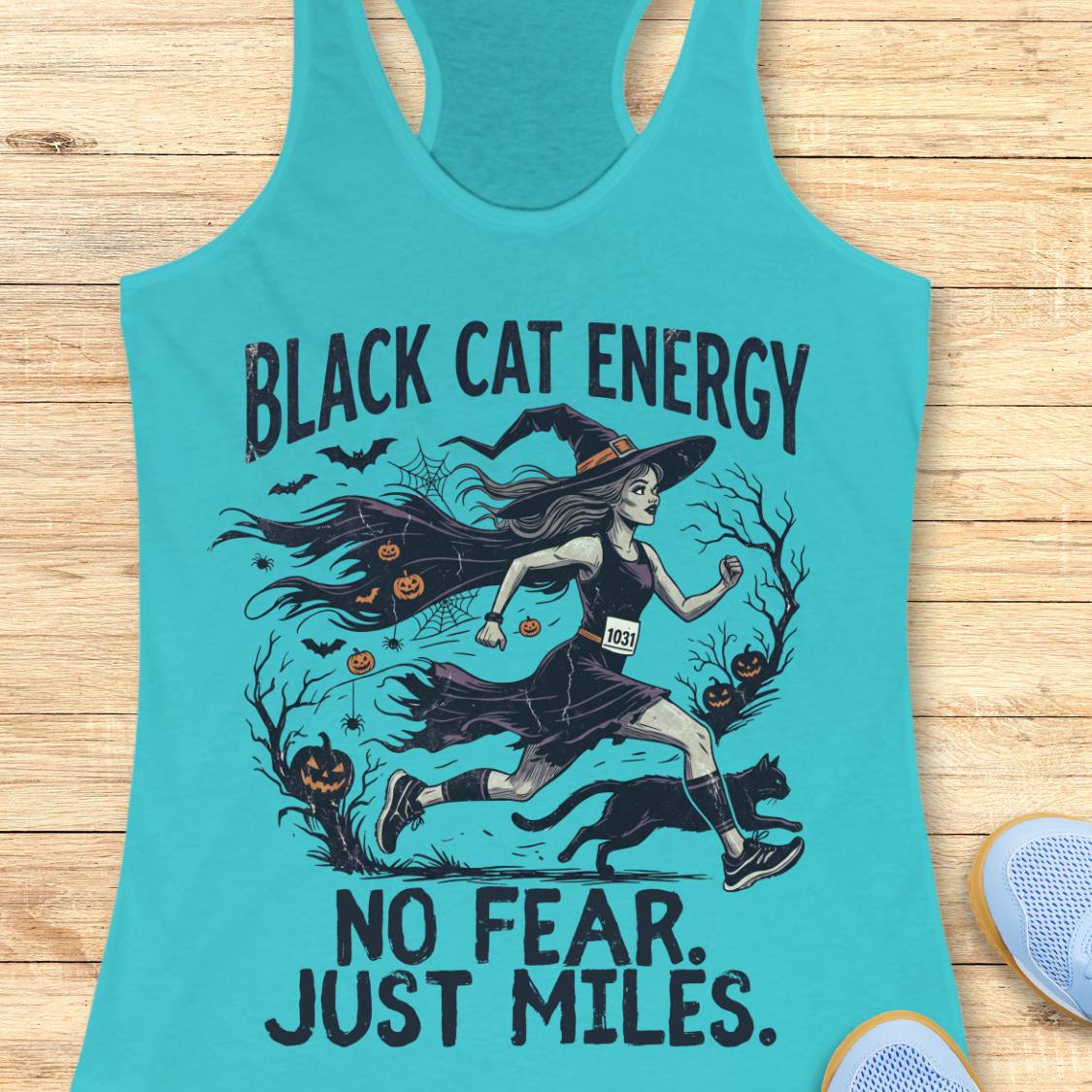 A turquoise racerback tank top laid flat on a light wood surface, featuring a Halloween-themed graphic of a witch running with a black cat and the text 'BLACK CAT ENERGY NO FEAR. JUST MILES.