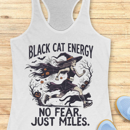 A white racerback tank top laid flat on a light wood surface, featuring a Halloween graphic of a witch running with a black cat and the text 'BLACK CAT ENERGY NO FEAR. JUST MILES.