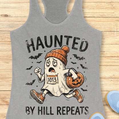 A heather gray racerback tank top laid flat on a light wood surface, featuring a Halloween graphic with the text 'HAUNTED BY HILL REPEATS' and an illustrated ghost running while carrying a pumpkin and wearing an orange beanie.