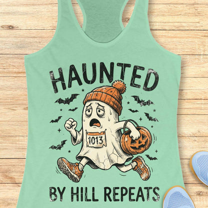 A mint green racerback tank top laid flat on a light wood background, featuring a cartoon ghost runner graphic with the text 'HAUNTED BY HILL REPEATS' and a pumpkin bucket.