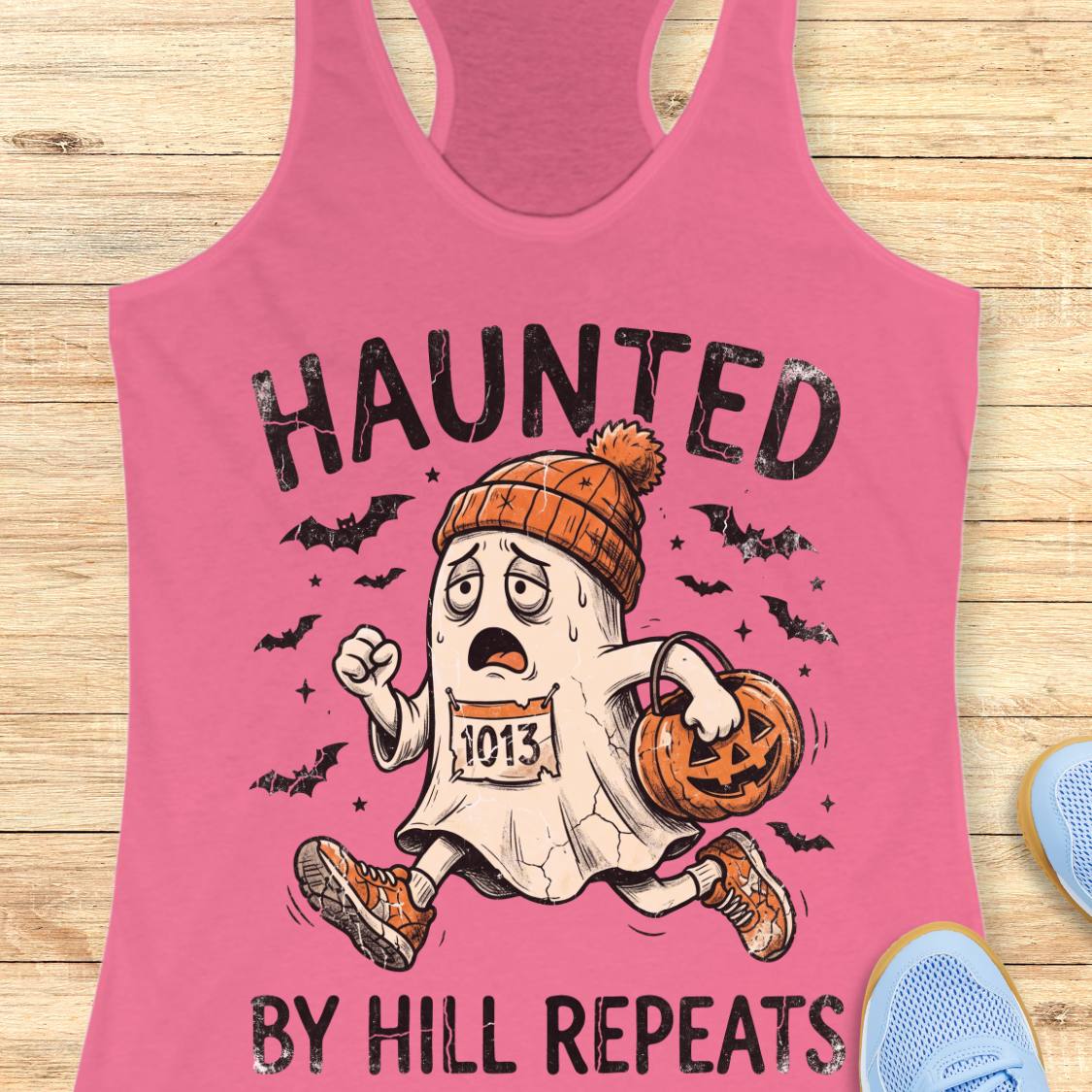 A pink racerback tank top laid flat on a light wood surface, featuring a Halloween graphic of a ghost running with a pumpkin bucket and the text 'HAUNTED BY HILL REPEATS.' Partial blue shoes are visible at the edges as styling props.
