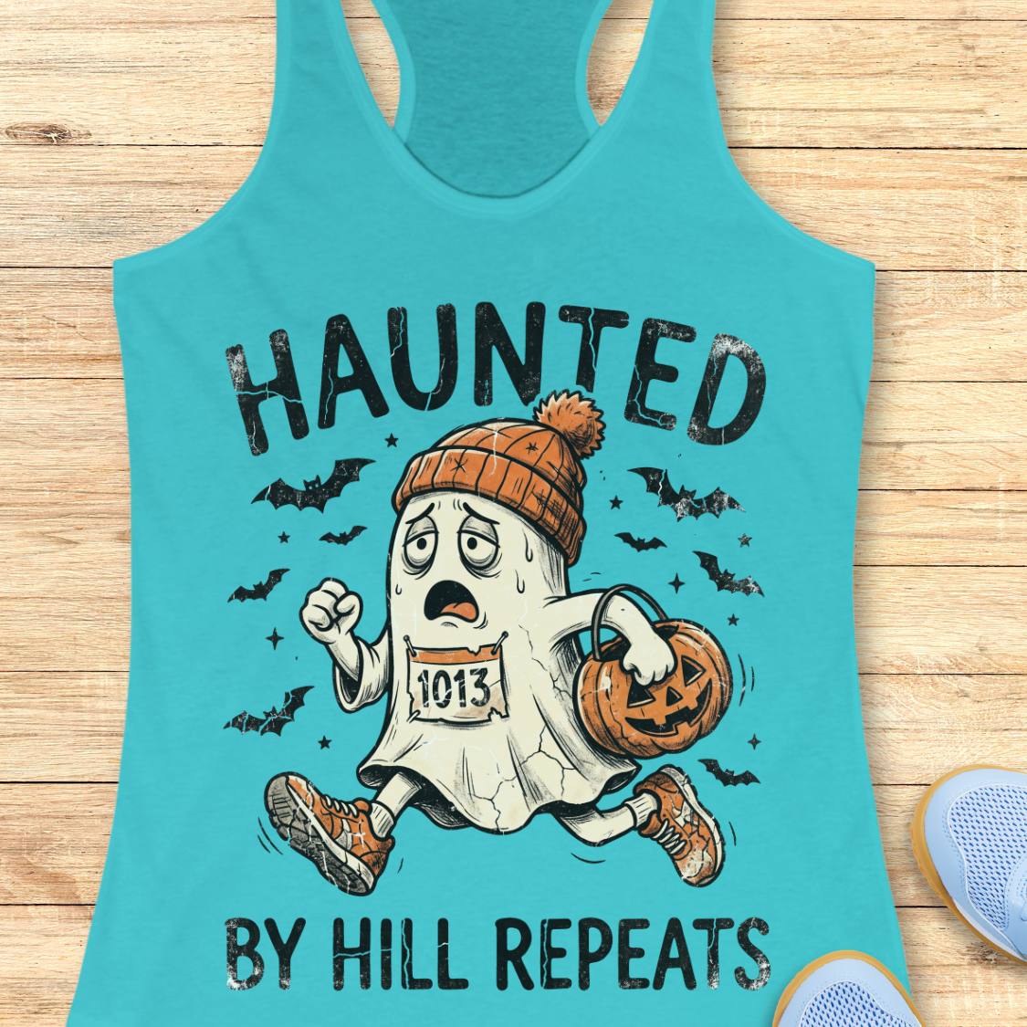 A turquoise racerback tank top laid flat on a light wood surface, featuring a Halloween graphic of a ghost running with a pumpkin and the text 'HAUNTED BY HILL REPEATS'.
