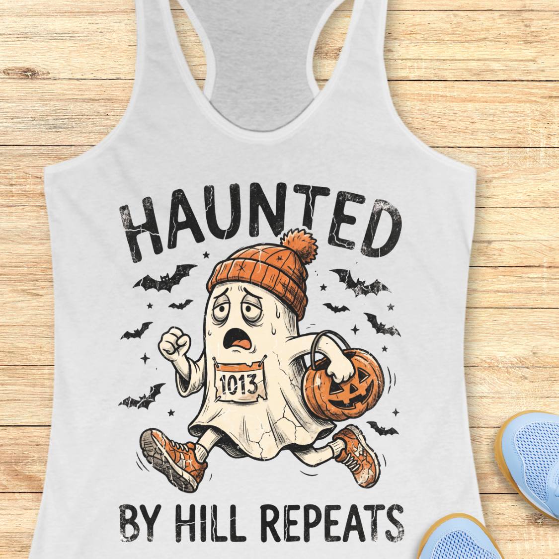 A white racerback tank top laid flat on a light wood background, featuring a Halloween-themed graphic with the text 'HAUNTED BY HILL REPEATS' and an illustrated ghost runner wearing an orange beanie and holding a pumpkin bucket.