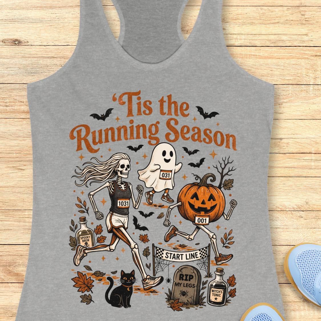 A heather gray racerback tank top laid flat on a light wood surface, featuring a Halloween-themed running graphic with skeleton, ghost, pumpkin, bats, leaves, and the text “Tis the Running Season.”.