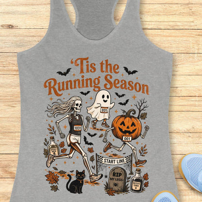 A heather gray racerback tank top laid flat on a light wood surface, featuring a Halloween-themed running graphic with skeleton, ghost, pumpkin, bats, leaves, and the text “Tis the Running Season.”.