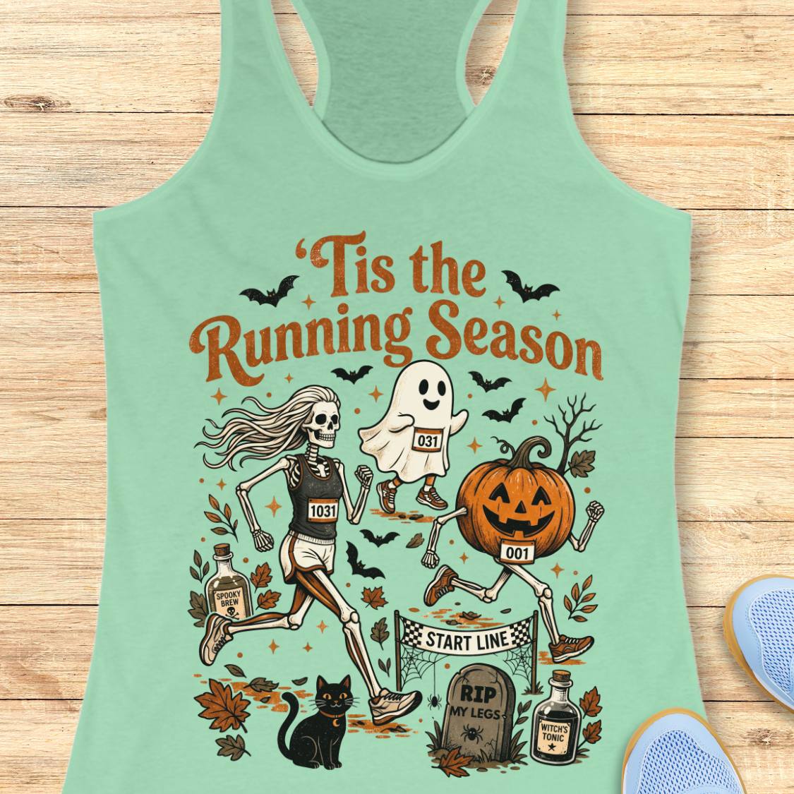 A mint green racerback tank top laid flat on a light wood background, featuring a colorful Halloween-themed graphic with the text 'Tis the Running Season' and illustrations of a skeleton runner, a ghost, a pumpkin, bats, a cat, and tombstones.