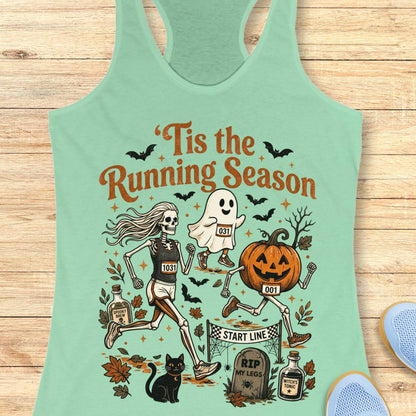 A mint green racerback tank top laid flat on a light wood background, featuring a colorful Halloween-themed graphic with the text 'Tis the Running Season' and illustrations of a skeleton runner, a ghost, a pumpkin, bats, a cat, and tombstones.