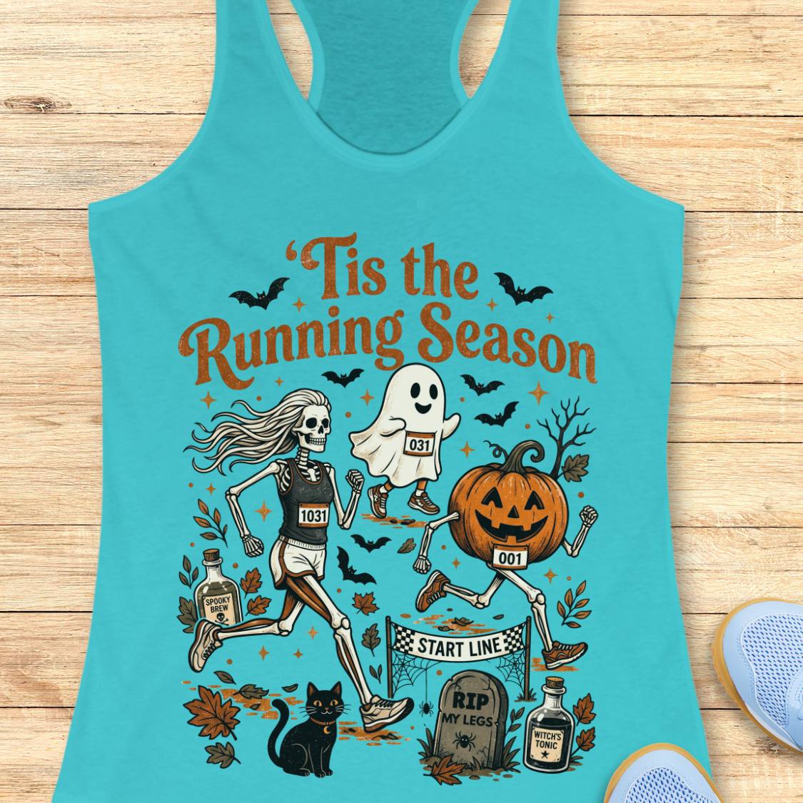 A turquoise racerback tank top laid flat on a light wood surface, featuring a Halloween-themed running graphic with skeleton, ghost, pumpkin, bats, leaves, and the text 'Tis the Running Season.' Partial blue sneakers are visible at the lower right edge.