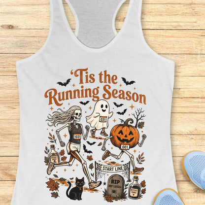 A white racerback tank top laid flat on a light wood surface, featuring a colorful Halloween-themed graphic with skeletons, a ghost, a jack-o'-lantern, bats, leaves, and the text “Tis the Running Season.”.