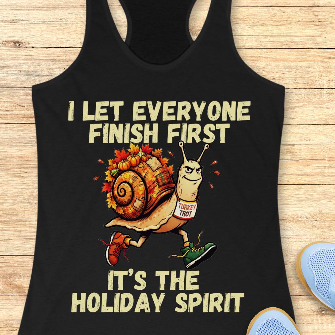 A black racerback tank top laid flat on a light wood surface, featuring a cartoon snail wearing a 'TURkey TROT' sign and running shoes, with the text 'I LET EVERYONE FINISH FIRST' and 'IT'S THE HOLIDAY SPIRIT'.
