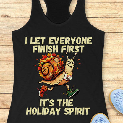 A black racerback tank top laid flat on a light wood surface, featuring a cartoon snail wearing a 'TURkey TROT' sign and running shoes, with the text 'I LET EVERYONE FINISH FIRST' and 'IT'S THE HOLIDAY SPIRIT'.