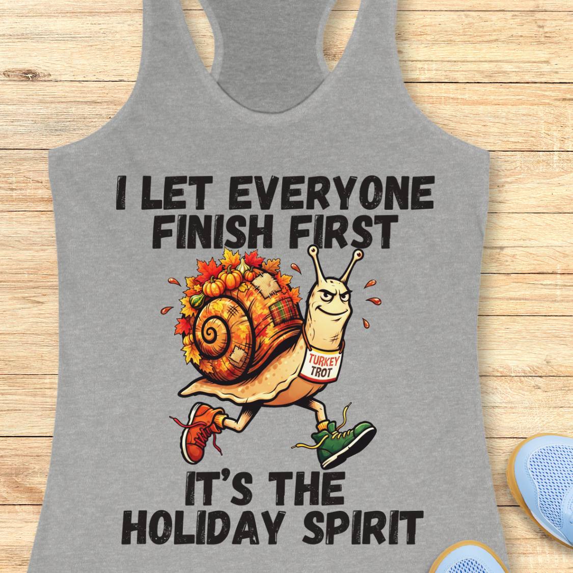 A heather gray racerback tank top laid flat on a light wood surface, featuring a holiday-themed graphic of a snail runner with the text 'I LET EVERYONE FINISH FIRST IT'S THE HOLIDAY SPIRIT'.