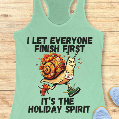 A mint green racerback tank top laid flat on a light wood background, featuring a holiday-themed graphic of a snail wearing a 'Turkey trot' sign and the text 'I LET EVERYONE FINISH FIRST IT'S THE HOLIDAY SPIRIT'.