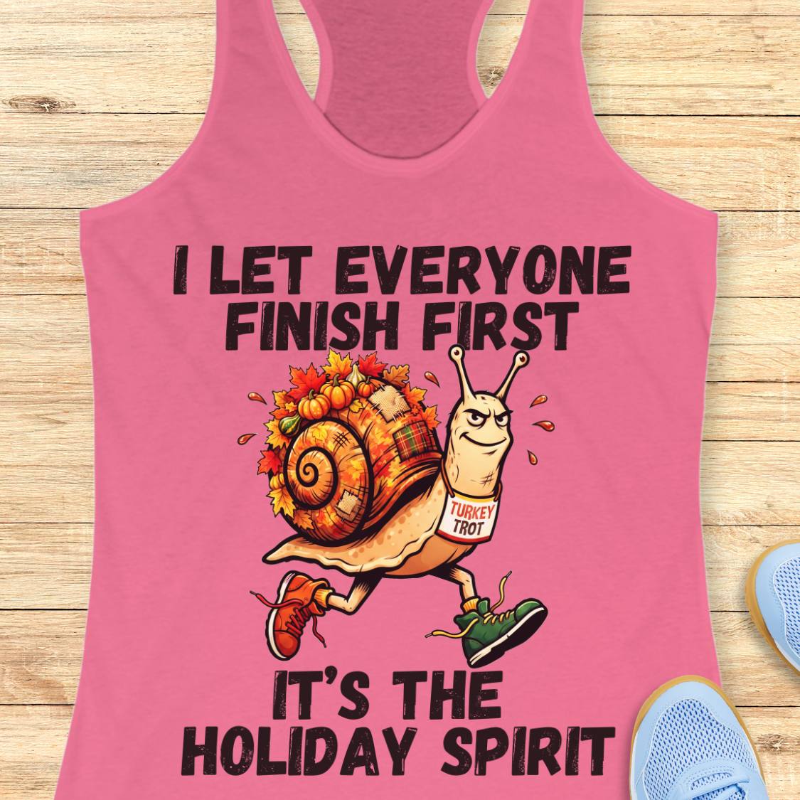 A pink racerback tank top laid flat on a light wood surface, featuring a cartoon snail wearing a 'Turkey Trot' bib and the text 'I LET EVERYONE FINISH FIRST IT'S THE HOLIDAY SPIRIT.' Partial blue shoes are visible at the edges as styling props.