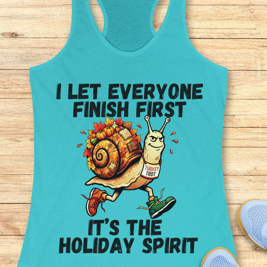 A turquoise racerback tank top laid flat on a light wood surface, featuring a holiday-themed graphic of a snail wearing a 'TUrkey trot' sign and the text 'I LET EVERYONE FINISH FIRST IT'S THE HOLIDAY SPIRIT'.