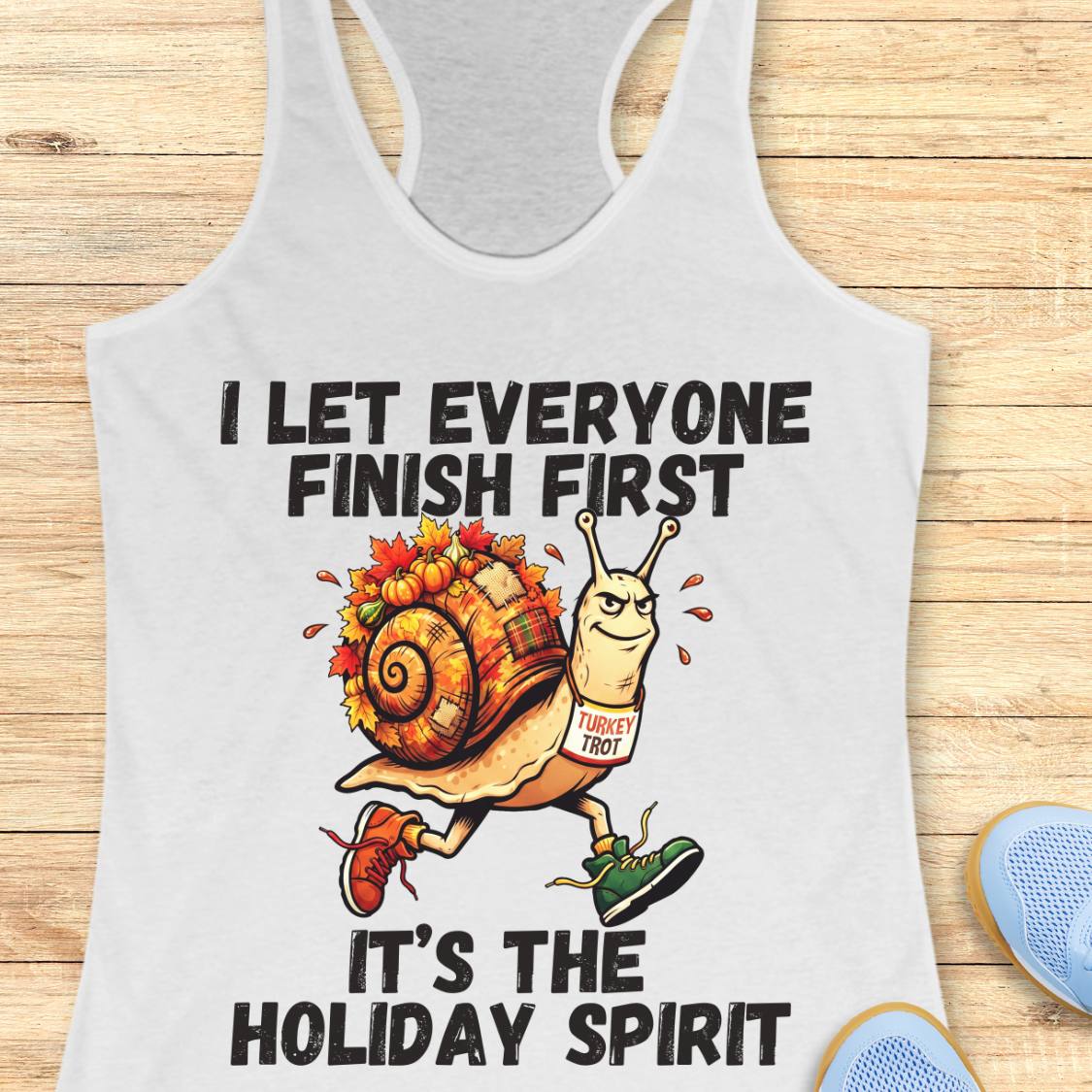 A white racerback tank top laid flat on a light wood surface, featuring a holiday-themed graphic of a snail wearing a Target logo shirt and colorful autumn leaves on its shell, with the text 'I LET EVERYONE FINISH FIRST' and 'IT'S THE HOLIDAY SPIRIT'. Partial blue shoes are visible at the bottom right as styling props.