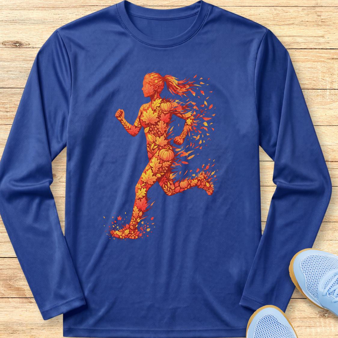 A royal blue long-sleeve athletic t-shirt laid flat on a light wood background, featuring a vibrant graphic of a running woman made of orange and yellow splatter-like shapes. A pair of light blue athletic shoes is partially visible in the bottom right corner.
