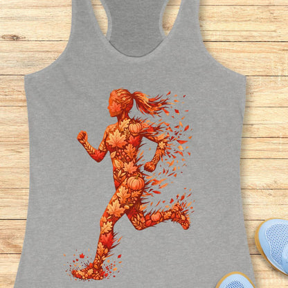 A heather gray racerback tank top featuring a graphic of a female runner composed of autumn leaves, pumpkins, and orange splatters, laid flat on a light wood plank background.