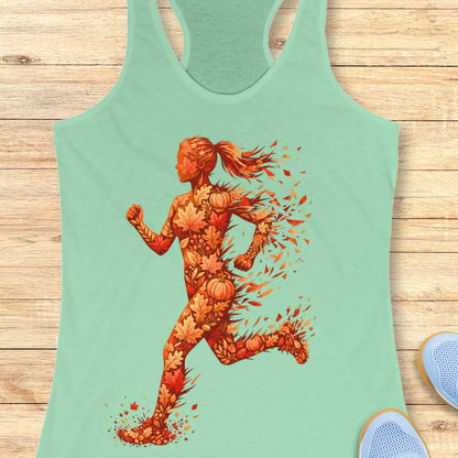 A mint green racerback tank top laid flat on a light wood background, featuring a large graphic of a running female silhouette composed of autumn leaves, pumpkins, and orange splatters.