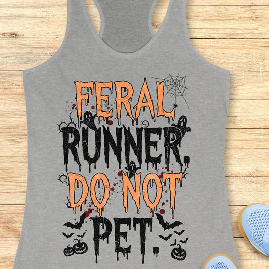 A heather gray racerback tank top with a Halloween-themed graphic reading 'FERAL RUNNER, DO NOT PET' in orange and black dripping lettering, styled flat on a light wood background with partial blue shoes visible at the edges.