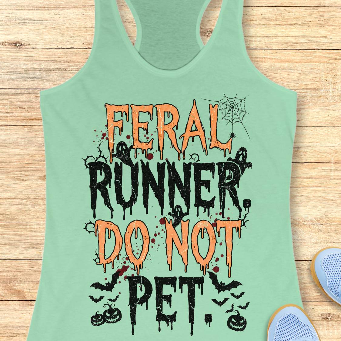 A mint green racerback tank top with a Halloween-themed graphic reading 'FERAL RUNNER, DO NOT PET.' in orange and black dripping lettering, styled flat on a light wood plank background with partial blue shoes visible at the edges.