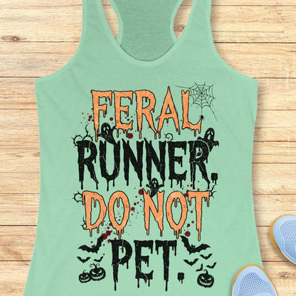 A mint green racerback tank top with a Halloween-themed graphic reading 'FERAL RUNNER, DO NOT PET.' in orange and black dripping lettering, styled flat on a light wood plank background with partial blue shoes visible at the edges.