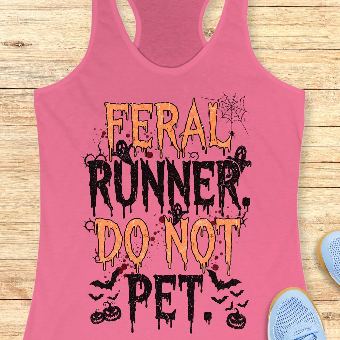 A pink racerback tank top laid flat on a light wood surface, featuring a Halloween-style graphic that reads “FERAL RUNNER, DO NOT PET.” in orange and black dripping lettering with spiderwebs, bats, and pumpkins.