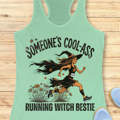 A mint green racerback tank top laid flat on a light wood background, featuring a graphic of a woman running as a witch with the text 'SOMEONE'S COOL-ASS RUNNING WITCH BESTIE'.